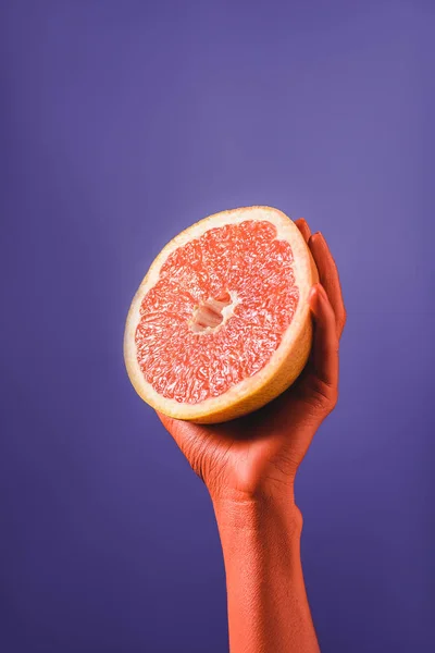 Vista recortada de mujer sosteniendo toronja cortada en mano de color coral sobre fondo azul sobre fondo violeta, concepto de color de 2019 - foto de stock