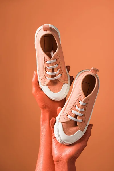 Partial view of woman holding sneakers on coral background, color of 2019 concept — Stock Photo