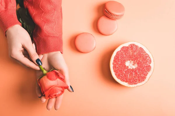 Cropped view of woman holding coral rose flower, macarons and grapefruit half on coral background, color of 2019 concept — Stock Photo