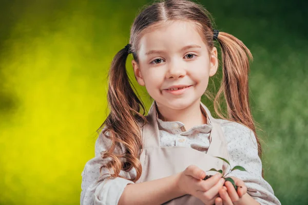 Улыбающийся ребенок держит молодое зеленое растение на размытом фоне, концепция Дня Земли — стоковое фото