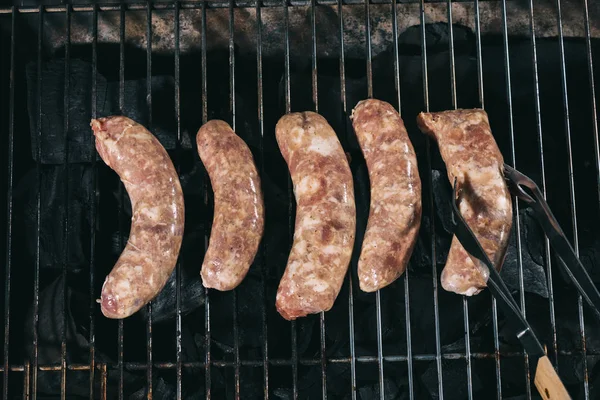 Top view of tweezers near uncooked fresh sausages on bbq grill grates — Stock Photo