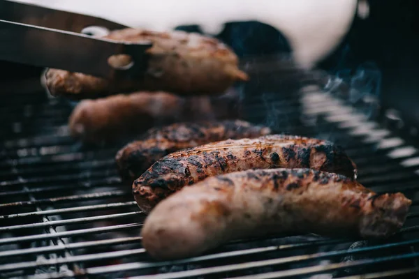 Selective focus of grilled tasty meat sausages and tweezers on barbecue grid — Stock Photo