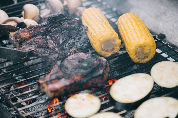 Selective focus of tweezers and juicy tasty steaks grilling on bbq grid with mushrooms, corn and sliced eggplant — Stock Photo