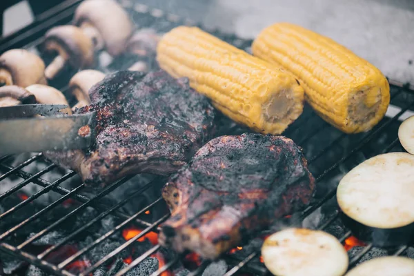 Fuoco selettivo di pinzette e succose gustose bistecche grigliate su griglia barbecue con funghi, mais e melanzane affettate — Foto stock
