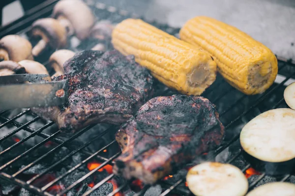 Selective focus of tweezers and juicy tasty steaks grilling on bbq grid with mushrooms, corn and sliced eggplant — Stock Photo