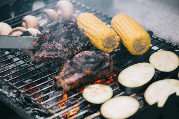 Selective focus of tweezers and juicy delicious steaks grilling on bbq grid with mushrooms, corn and sliced eggplant — Stock Photo