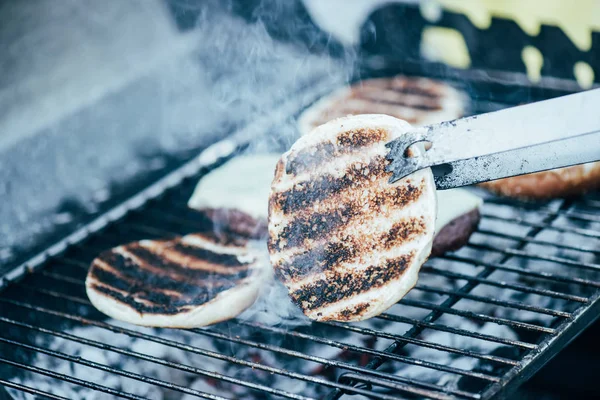Selective focus of tweezers and delicious fresh burgers ingredients with crust grilling on barbecue grid — Stock Photo