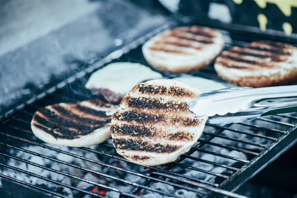 Selective focus of tweezers and delicious fresh burgers ingredients with crust grilling on barbecue grid — Stock Photo