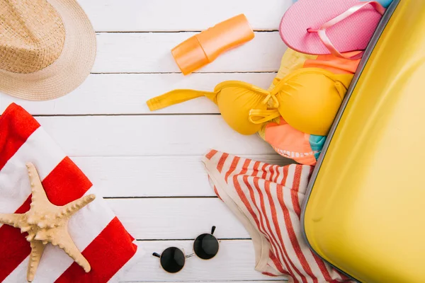 Vue de dessus de crème solaire, étoiles de mer et accessoires et vêtements d'été en valise sur fond de bois blanc — Photo de stock