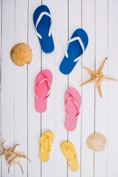 Top view of yellow, blue and pink flip flops on white wooden background with seashells — Stock Photo