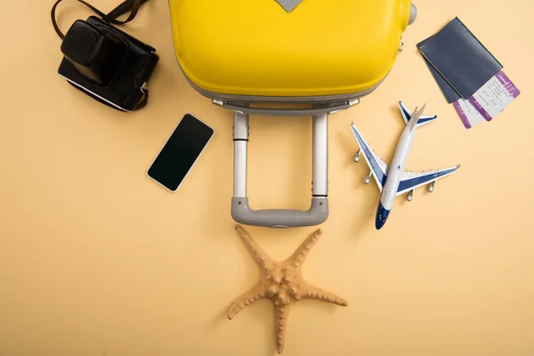 Top view of yellow suitcase, plane model, starfish, film camera, smartphone and tickets on beige background — Stock Photo