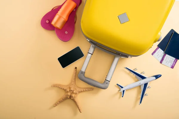 Vista superior de la maleta amarilla, modelo de avión, estrella de mar, protector solar, chanclas, teléfono inteligente y boletos sobre fondo beige - foto de stock
