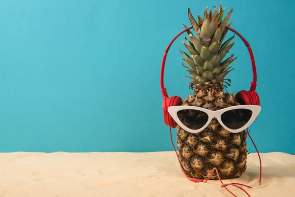 Ananas mit Kopfhörern und Sonnenbrille auf Sand auf blauem Hintergrund — Stockfoto