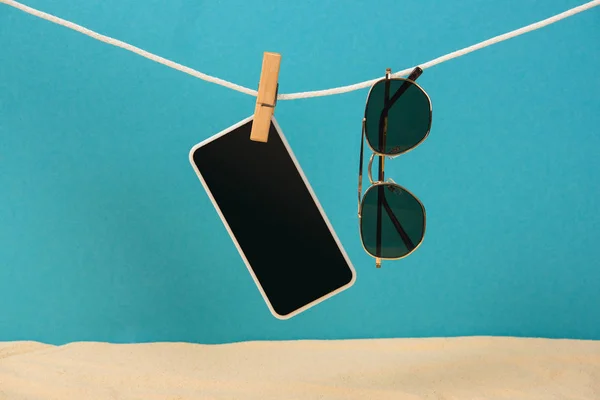 Smartphone with blank screen and sunglasses hanging on rope with pin on blue background — Stock Photo