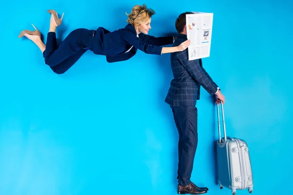 Femme posant derrière l'homme avec une valise et tenant un journal près de son visage sur fond bleu — Photo de stock