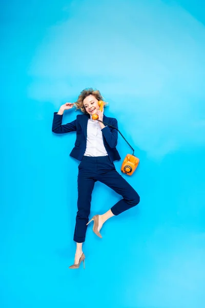 Mujer elegante en traje y zapatos de tacón alto hablando por teléfono sobre fondo azul - foto de stock