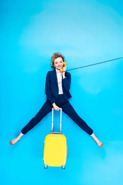 Encantadora mujer de traje con maleta amarilla hablando por teléfono sobre fondo azul - foto de stock
