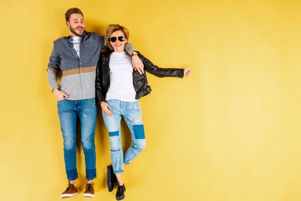 Mujer sonriente mostrando el pulgar hacia arriba y abrazándose con el novio sobre fondo amarillo - foto de stock