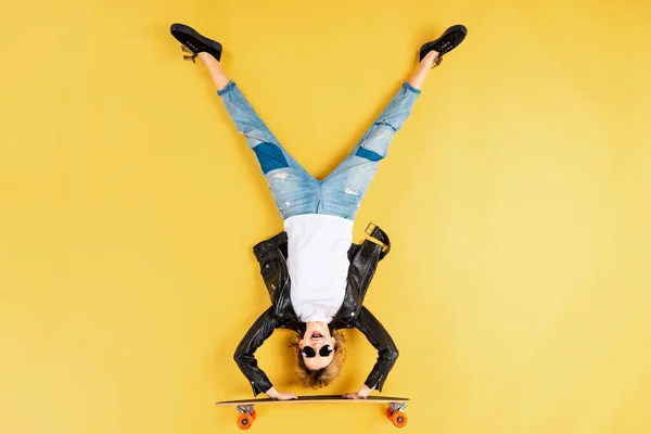 Girl in leather jacket standing on hands on longboard on yellow background — Stock Photo