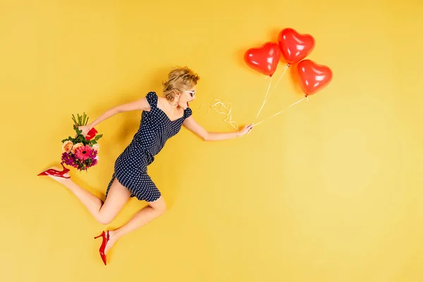 Femme excitée avec des ballons à air en forme de coeur et des fleurs sautant sur fond jaune — Photo de stock