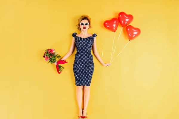 Mulher elegante segurando balões de ar em forma de coração e flores no fundo amarelo — Fotografia de Stock