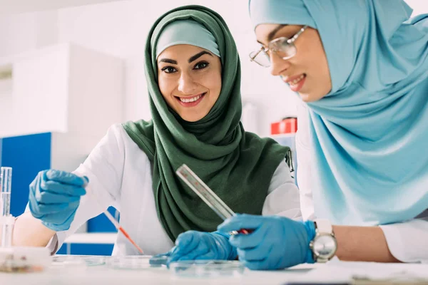 Des scientifiques musulmanes souriantes tenant une pipette et un tube à essai en verre lors d'une expérience en laboratoire chimique — Photo de stock