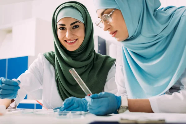 Científicos musulmanes hembras sosteniendo pipeta y tubo de ensayo de vidrio durante el experimento en laboratorio químico - foto de stock