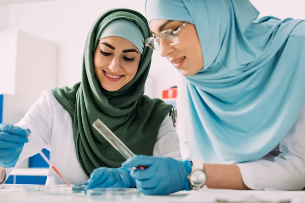 Des scientifiques musulmanes souriantes tenant une pipette et un tube à essai en verre tout en expérimentant en laboratoire chimique — Photo de stock