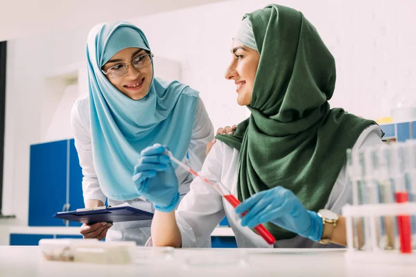 Sonrientes científicas musulmanas en hijab con tubo de ensayo y pipeta durante el experimento en laboratorio químico - foto de stock