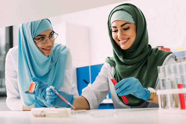 Científicos musulmanes sonrientes en hijab con tubo de ensayo y pipeta durante el experimento en laboratorio químico - foto de stock
