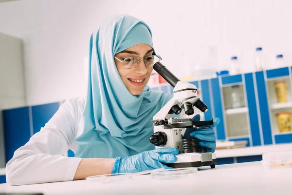 Científica musulmana sonriente mirando a través del microscopio durante el experimento en laboratorio químico - foto de stock