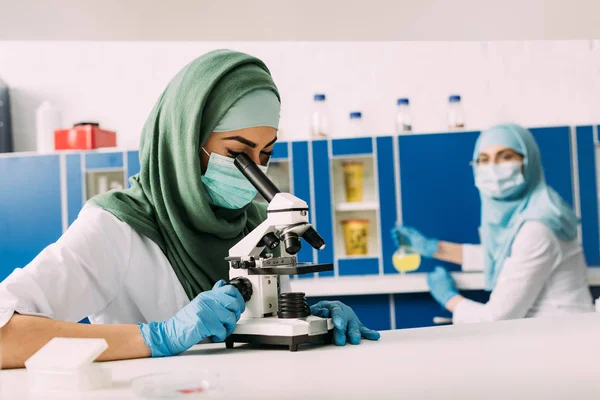 Científica musulmana hembra mirando a través del microscopio durante el experimento con un colega que trabaja en el fondo en el laboratorio químico - foto de stock