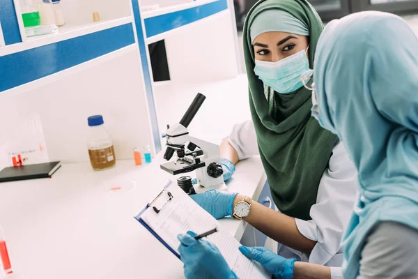 Científicas musulmanas en hijab utilizando microscopio y portapapeles durante el experimento en laboratorio químico - foto de stock