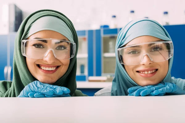 Улыбающиеся мусульманские химики в защитных очках и хиджабе, смотрящие в камеру в лаборатории — стоковое фото