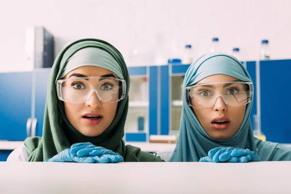 Mulheres químicas muçulmanas chocadas em óculos de proteção e hijab olhando para a câmera em laboratório — Fotografia de Stock