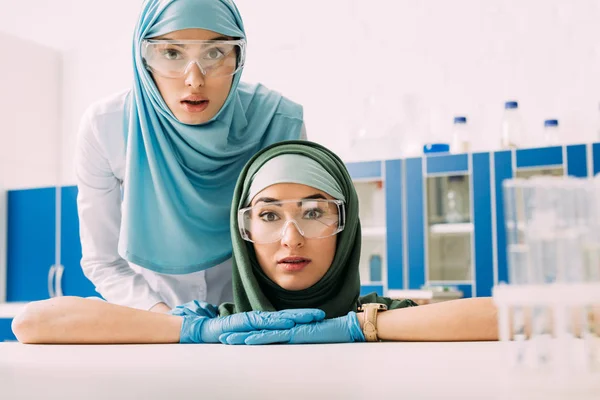 Female muslim chemists in protective goggles and hijab looking at camera in laboratory — Stock Photo