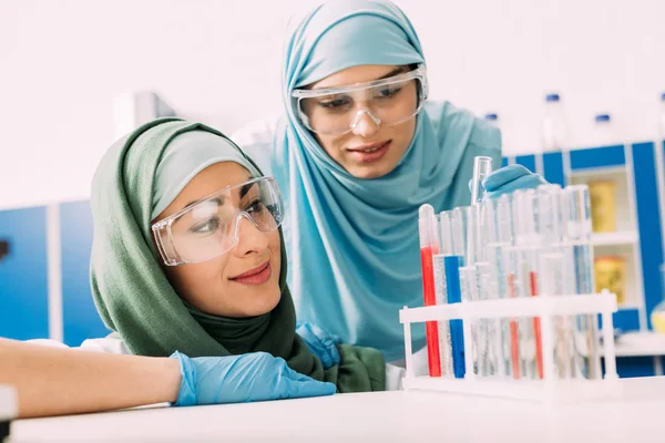 Científicas musulmanas en gafas que observan reactivos en tubos de ensayo de vidrio durante el experimento en laboratorio químico - foto de stock