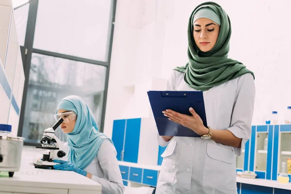 Mulheres cientistas muçulmanas concentradas usando microscópio e escrita em prancheta durante experimento em laboratório químico — Fotografia de Stock