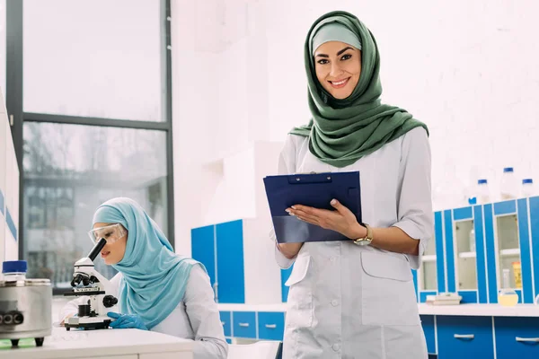 Científicos musulmanes femeninos que utilizan microscopio y portapapeles durante el experimento en laboratorio químico - foto de stock