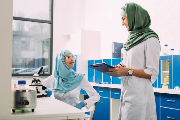 Científicos musulmanes femeninos que utilizan microscopio y portapapeles durante el experimento en laboratorio químico - foto de stock