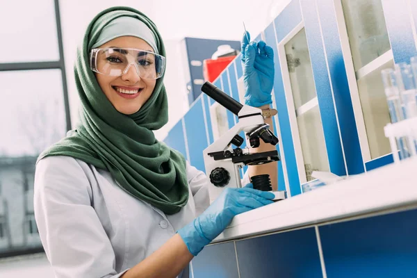 Muslimische Wissenschaftlerin sitzt mit Mikroskop am Tisch, hält Glasprobe und blickt in Chemielabor in die Kamera — Stockfoto