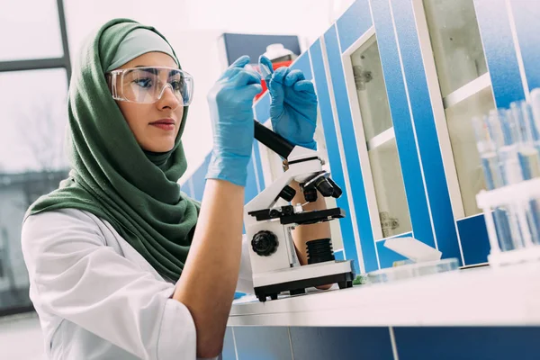 Female muslim scientist sitting at table with microscope and looking at glass sample during experiment in chemical laboratory — Stock Photo