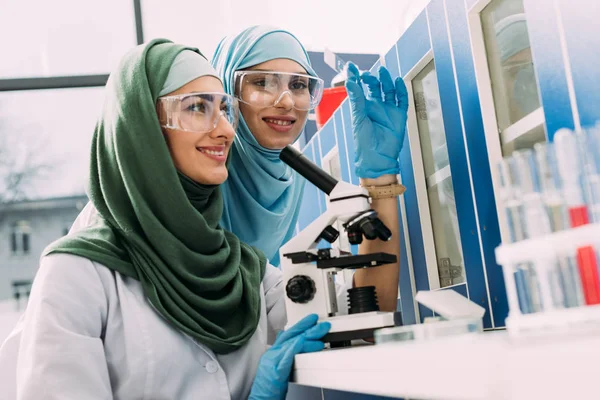 Mulheres cientistas muçulmanas sorridentes com microscópio e amostra de vidro durante o experimento em laboratório químico — Fotografia de Stock