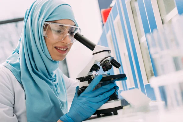 Científica musulmana sonriente mirando a través del microscopio durante el experimento en laboratorio químico - foto de stock