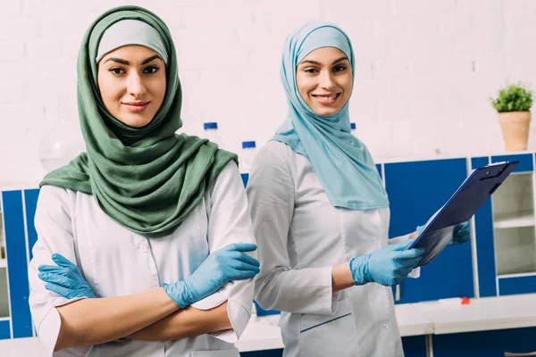 Mulheres químicas muçulmanas bonitas em hijab com braços cruzados e prancheta olhando para a câmera em laboratório — Fotografia de Stock