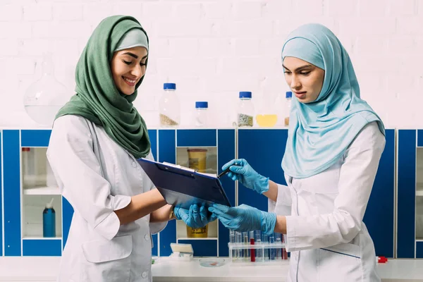 Femmes musulmanes scientifiques dans le hijab tenant presse-papiers pendant l'expérience en laboratoire chimique — Photo de stock
