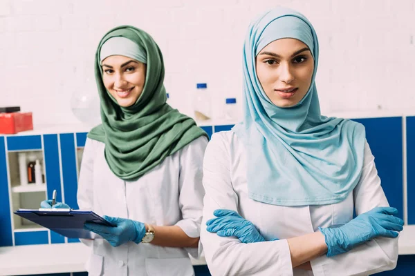 Mulheres químicas muçulmanas bonitas em hijab com braços cruzados e prancheta olhando para a câmera em laboratório — Fotografia de Stock