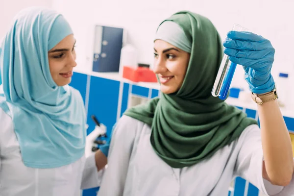 Улыбающиеся мусульманские ученые держат стеклянные пробирки с реагентами во время эксперимента в химической лаборатории — стоковое фото