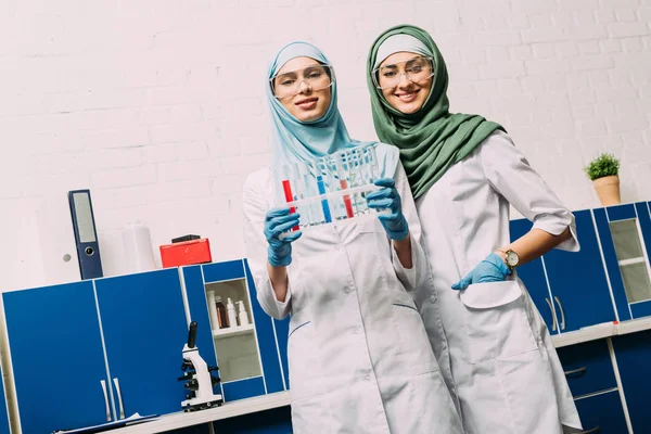 Жінки-мусульмани з пробірками, які дивляться на камеру під час експерименту в хімічній лабораторії — стокове фото