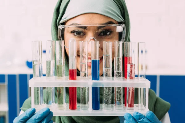 Sonriente mujer musulmana científica sosteniendo tubos de ensayo con líquido rojo y azul mientras mira a la cámara - foto de stock